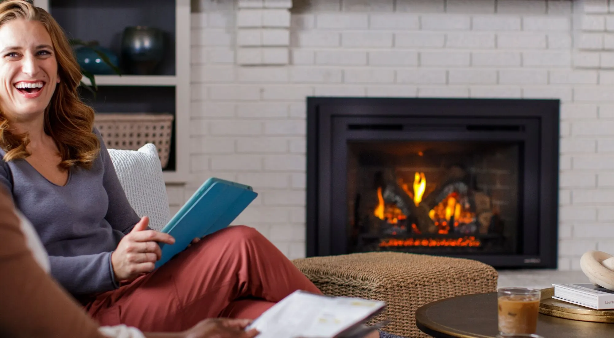 A couple laughing together in their living room beside a lit gas insert in a white brick fireplace