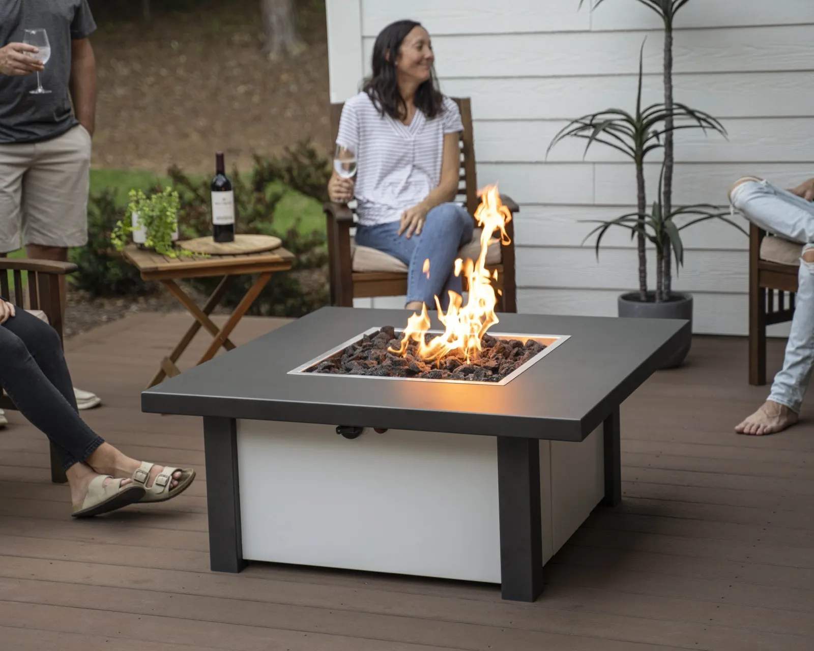 Friends sharing wine around a lit Stoll fire pit table on a backyard deck at dusk