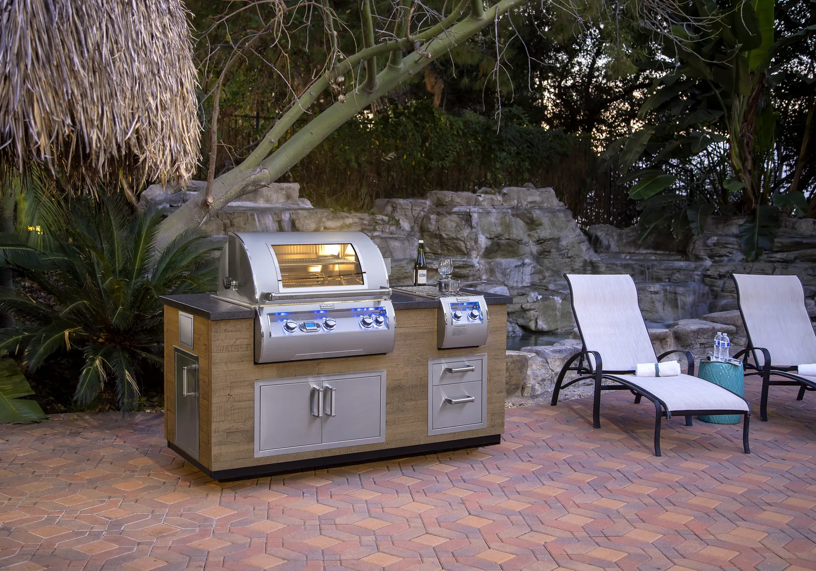 A lit Napoleon Echelon grill in a wood-paneled island with stainless doors and drawers on a poolside patio at dusk